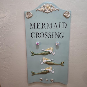 May include: A light blue wooden sign with the words "MERMAID CROSSING" in black. The sign features three mermaid figures with gold tails and blonde hair. Decorative seashells and jewels adorn the sign.