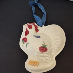 May include: A white ceramic rooster ornament with red comb and wattle, and red and pink floral accents. The rooster has a blue ribbon for hanging. The ornament is a decorative item.