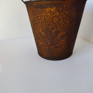 May include: A small, rusty brown metal bucket with a handle. The bucket has the words "SWEET CORN" embossed on the front, along with a leaf design. The bucket is likely for decorative use.