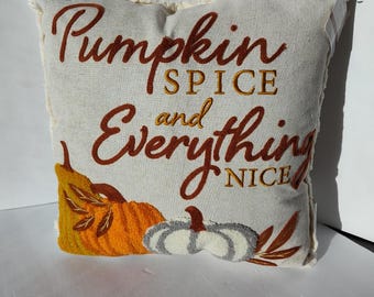 May include: Square decorative pillow with embroidered text "Pumpkin Spice and Everything Nice". The pillow features embroidered pumpkins in orange and gray, with brown leaves. The pillow has a cream-colored background and a fringed edge.