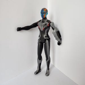 Op de afbeelding: Een plastic actiefiguur van een personage met een blauwe, oranje en zwarte huid. De figuur draagt een zwart en zilver pak met het Avengers-logo op de borst. De figuur poseert met &eacute;&eacute;n arm uitgestrekt.