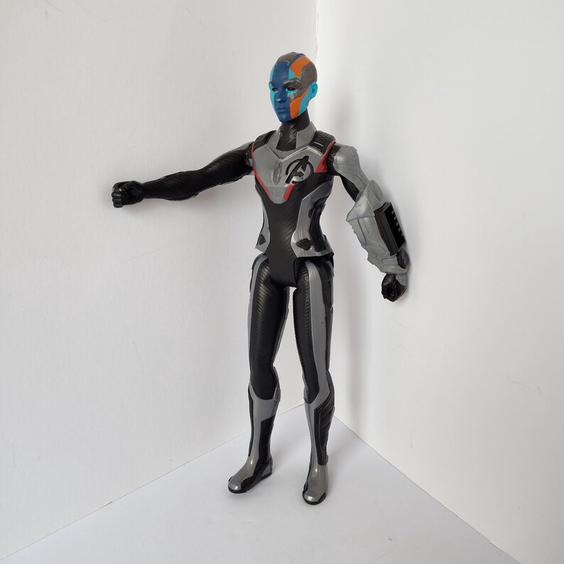 Op de afbeelding: Een plastic actiefiguur van een personage met een blauwe, oranje en zwarte huid. De figuur draagt een zwart en zilver pak met het Avengers-logo op de borst. De figuur poseert met &eacute;&eacute;n arm uitgestrekt.