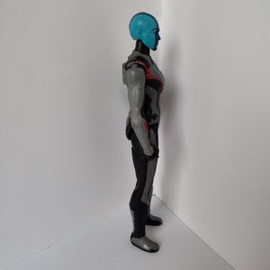 Op de afbeelding: Zijaanzicht van een blauwhuidige actiefiguur met een blauwe helm en een grijs en zwart pak. De figuur heeft rode accenten op de schouders en armen. De figuur staat op een wit oppervlak.