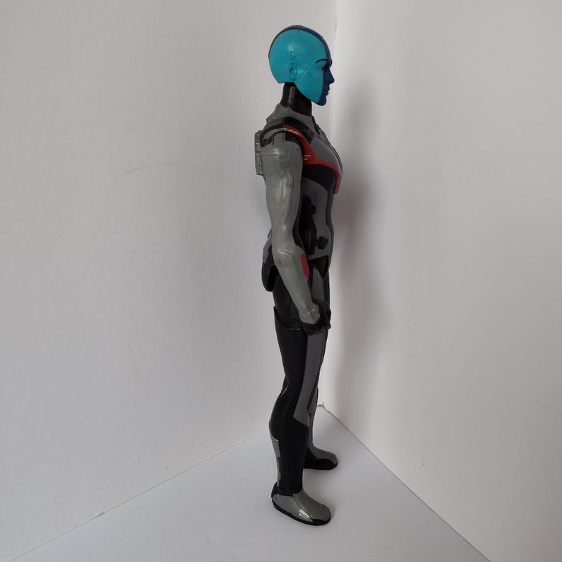 Op de afbeelding: Zijaanzicht van een blauwhuidige actiefiguur met een blauwe helm en een grijs en zwart pak. De figuur heeft rode accenten op de schouders en armen. De figuur staat op een wit oppervlak.