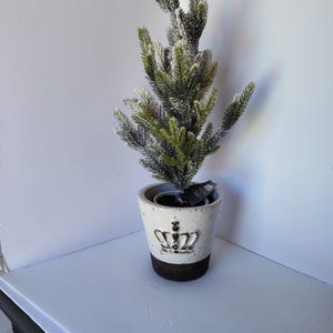 May include: A small, artificial evergreen tree in a white ceramic pot with a brown base and a crown design. The tree has green needles dusted with white, creating a snowy effect. The pot is on a white surface.