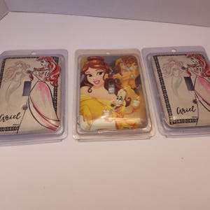 Pu&ograve; includere: Tre copri interruttori a tema Disney in confezione di plastica. Uno presenta Ariel, un altro Belle e il terzo di nuovo Ariel. Le copertine di Ariel la mostrano con un vestito rosa. La copertina di Belle la mostra con un vestito giallo con Lumi&egrave;re.