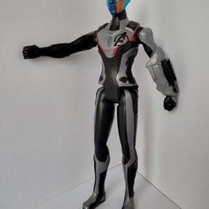 Op de afbeelding: Een plastic actiefiguur van een personage met blauwe, oranje en zwarte gezichtsverf. De figuur draagt een zwart en grijs pak met het Avengers-logo op de borst. De figuur poseert met &eacute;&eacute;n arm uitgestrekt.