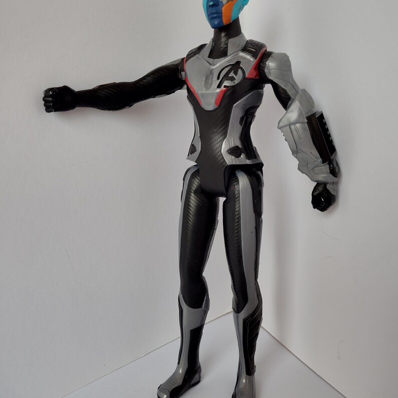 Op de afbeelding: Een plastic actiefiguur van een personage met blauwe, oranje en zwarte gezichtsverf. De figuur draagt een zwart en grijs pak met het Avengers-logo op de borst. De figuur poseert met &eacute;&eacute;n arm uitgestrekt.