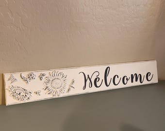 May include: A rectangular, off-white wooden sign with the word "Welcome" in black script. The left side features embossed floral and insect designs, including sunflowers, butterflies, and bees. The sign is a decorative home accent.