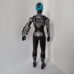 Op de afbeelding: Een plastic actiefiguur van een personage in een zwart en grijs pak met een blauw hoofd en een grijze arm. De figuur heeft een gedetailleerd ontwerp met een paneel op de rug en staat op een wit oppervlak.