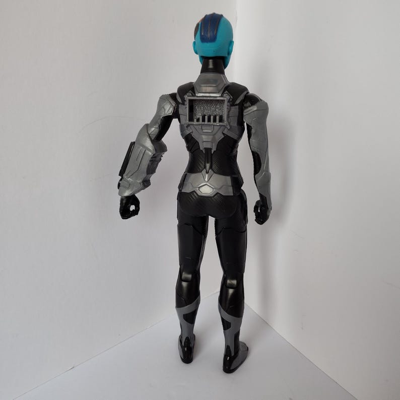 Op de afbeelding: Een plastic actiefiguur van een personage in een zwart en grijs pak met een blauw hoofd en een grijze arm. De figuur heeft een gedetailleerd ontwerp met een paneel op de rug en staat op een wit oppervlak.