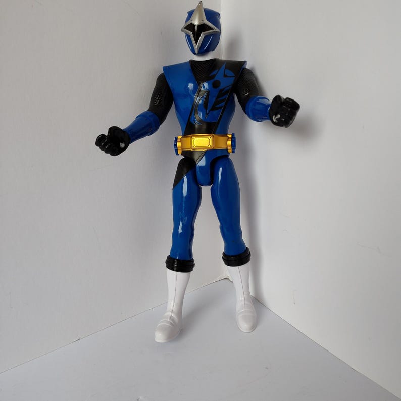 Op de afbeelding: Een blauw actiefiguur van een ninja-krijger met een zilveren helm en gouden riem. Het figuur heeft zwarte accenten op de armen en borst, witte laarzen en zwarte handschoenen. Het figuur staat in een neutrale pose.
