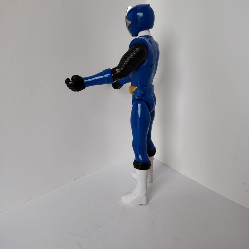 Op de afbeelding: Een blauw actiefiguur van een superheld in een glanzend blauw pak met zwarte accenten op de armen. Het figuur heeft een witte helm en laarzen en een gele riem. Het figuur poseert met uitgestrekte armen.