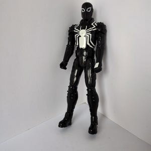 Op de afbeelding: Een zwart actiefiguur van een superheld met een wit spinnenembleem op de borst. Het figuur heeft een zwart masker en het pak is gedetailleerd met witte accenten. Het figuur staat in een neutrale pose.