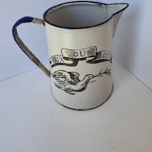 May include: Cream-colored enamel pitcher with a blue handle and rim. The pitcher features a black illustration of a dove carrying an olive branch, with the text "PAIX VOUS SOIT" in a banner above the dove.