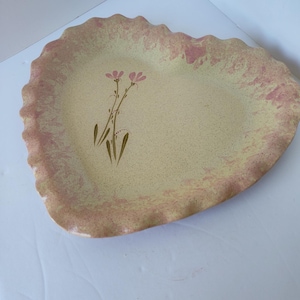 May include: A heart-shaped ceramic serving platter with a speckled beige center and a pink, scalloped edge. The platter features a floral design with pink flowers and green stems.