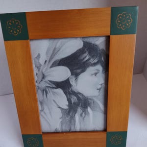 May include: Wooden picture frame with a portrait of a woman in black and white. The frame is light brown with dark green accents at the corners, each featuring a floral design. The portrait is a detailed drawing.