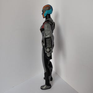 Op de afbeelding: Een zijaanzicht van een futuristisch actiefiguur. Het figuur is voornamelijk grijs en zwart met rode en blauwe accenten. Het heeft een helm met blauwe en oranje details en een gedetailleerd armpaneel.