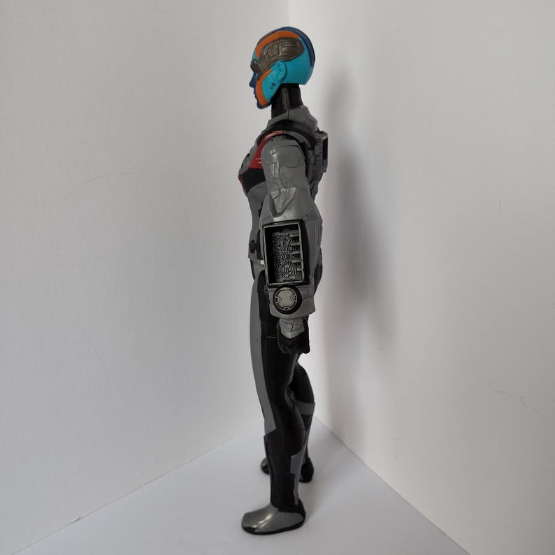 Op de afbeelding: Een zijaanzicht van een futuristisch actiefiguur. Het figuur is voornamelijk grijs en zwart met rode en blauwe accenten. Het heeft een helm met blauwe en oranje details en een gedetailleerd armpaneel.