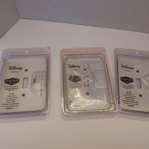 Pu&ograve; includere: Tre placche per interruttori Disney in confezione di plastica trasparente. Le placche sono bianche con un'apertura rettangolare e includono viti. La confezione presenta anche il marchio "Open Road" e informazioni sul prodotto.