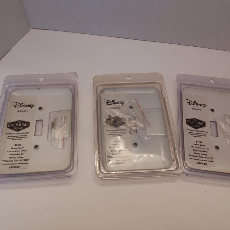 Pu&ograve; includere: Tre placche per interruttori Disney in confezione di plastica trasparente. Le placche sono bianche con un'apertura rettangolare e includono viti. La confezione presenta anche il marchio "Open Road" e informazioni sul prodotto.