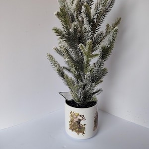 May include: A small artificial evergreen tree in a white ceramic pot. The tree has green needles dusted with white, and the pot features a badger illustration. The pot is approximately 4 inches tall.