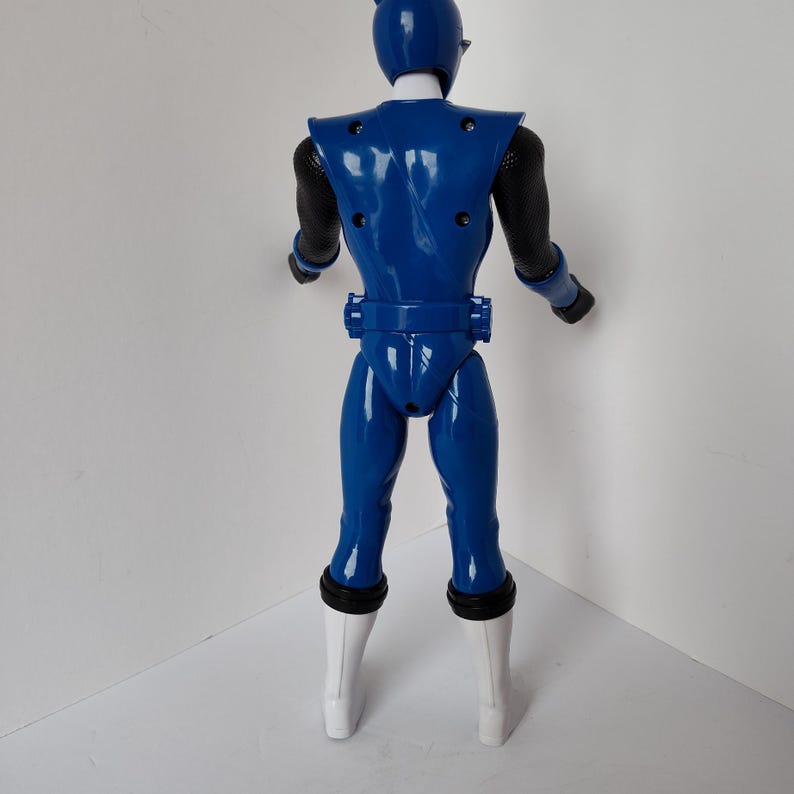 Op de afbeelding: Blauw actiefiguur van een superheld, van achteren gezien. De figuur heeft een blauwe helm, pak en witte laarzen. Zwarte mesh mouwen en handschoenen zijn zichtbaar. De figuur heeft een riem en een gestileerd ontwerp op de rug.