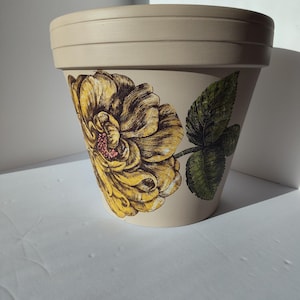 May include: A beige ceramic flower pot with a large yellow and brown floral design and green leaves. The pot has a wide rim and a slightly tapered shape. The floral design is detailed and appears to be hand-painted.