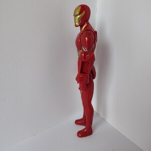 Op de afbeelding: Een rode Iron Man actiefiguur, in profiel. De figuur heeft een gouden helm met een geel vizier en gouden details op de borst en armen. De figuur is van plastic en staat tegen een witte achtergrond.
