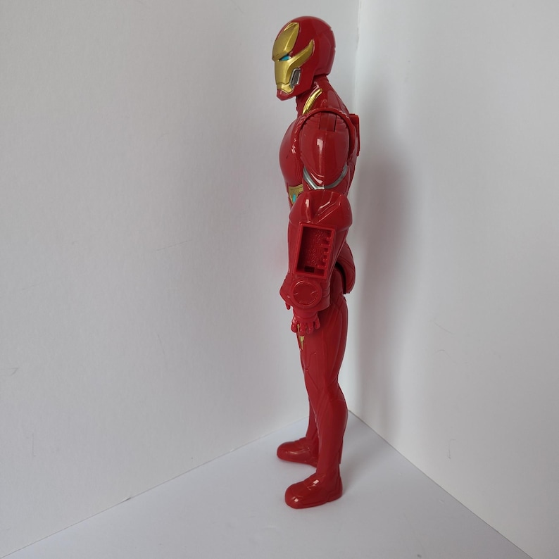 Op de afbeelding: Een rode Iron Man actiefiguur, in profiel. De figuur heeft een gouden helm met een geel vizier en gouden details op de borst en armen. De figuur is van plastic en staat tegen een witte achtergrond.