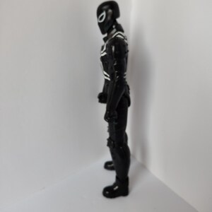 Op de afbeelding: Zwart actiefiguur van een superheld in een volledig pak met witte accenten. Het figuur heeft een helm met een wit oogontwerp. Het figuur staat in een neutrale houding.