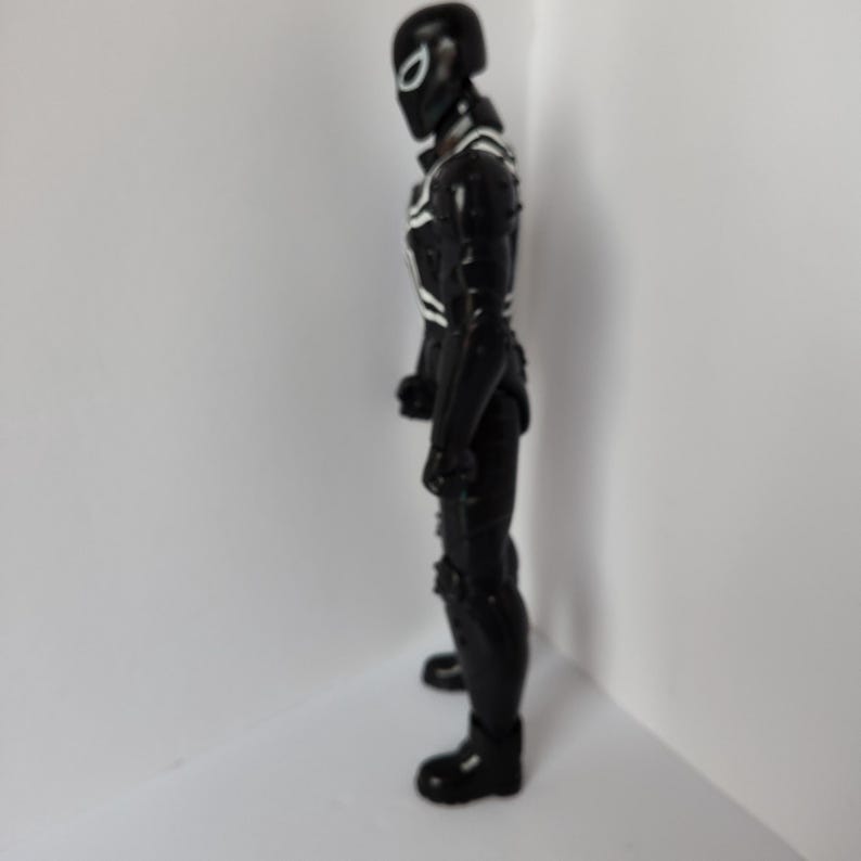 Op de afbeelding: Zwart actiefiguur van een superheld in een volledig pak met witte accenten. Het figuur heeft een helm met een wit oogontwerp. Het figuur staat in een neutrale houding.