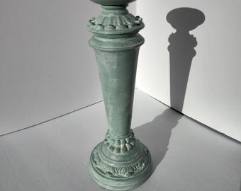 May include: A tall, sage green pedestal with a round top and ornate detailing. The pedestal has a flared, conical shape and decorative floral patterns at the base and midsection. The shadow of the pedestal is visible on the white background.