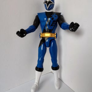 Op de afbeelding: Een blauw actiefiguur van een ninja-krijger met een zilveren helm en een gouden riem. Het figuur heeft zwarte handschoenen en witte laarzen. De ninja staat in een vechthouding.