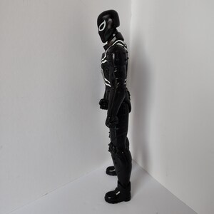 Op de afbeelding: Zwart actiefiguur van een superheld in een volledig pak. De figuur heeft een zwarte helm met een wit oogontwerp en witte accenten op de borst en armen. De figuur staat in een hoek.