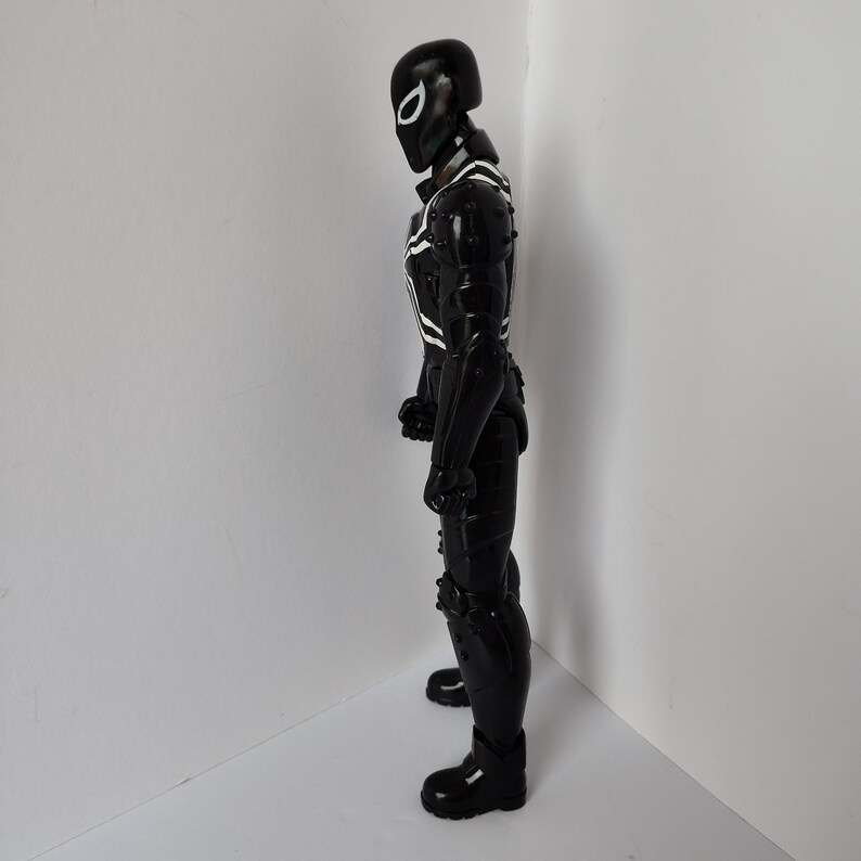 Op de afbeelding: Zwart actiefiguur van een superheld in een volledig pak. De figuur heeft een zwarte helm met een wit oogontwerp en witte accenten op de borst en armen. De figuur staat in een hoek.