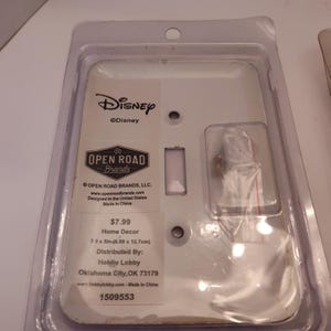 Pu&ograve; includere: Una placca interruttore bianca a tema Disney in una confezione di plastica trasparente. La placca ha un'apertura rettangolare per l'interruttore e due fori per le viti. La confezione include il logo Disney, il logo Open Road Brands e le informazioni sul prodotto.