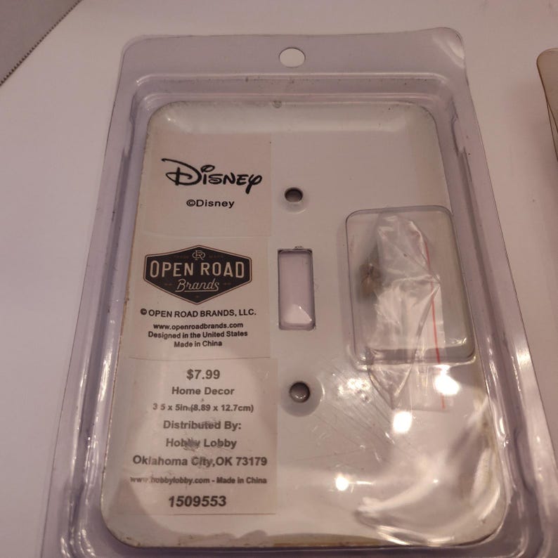 Pu&ograve; includere: Una placca interruttore bianca a tema Disney in una confezione di plastica trasparente. La placca ha un'apertura rettangolare per l'interruttore e due fori per le viti. La confezione include il logo Disney, il logo Open Road Brands e le informazioni sul prodotto.