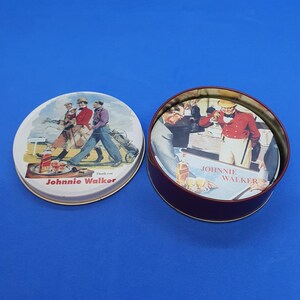 May include: A round, burgundy-colored tin with a lid featuring a vintage illustration of men playing golf. The lid is open, revealing a matching illustration inside. The text "Johnnie Walker" is visible.