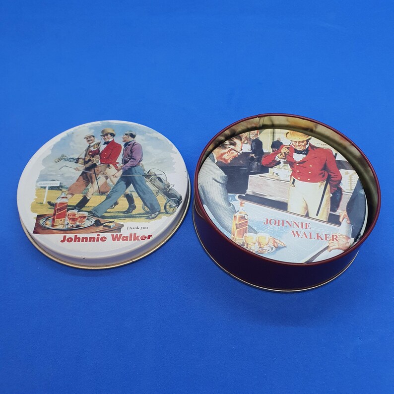 May include: A round, burgundy-colored tin with a lid featuring a vintage illustration of men playing golf. The lid is open, revealing a matching illustration inside. The text "Johnnie Walker" is visible.