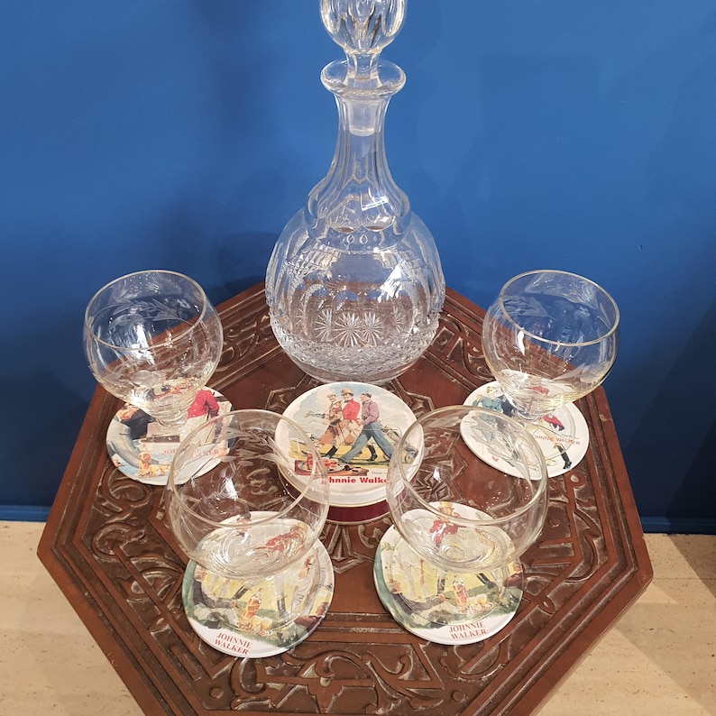May include: A crystal decanter with a stopper, surrounded by five clear, round glasses and coasters. The coasters feature a vintage illustration and the text "Johnnie Walker". The set is arranged on a wooden table.