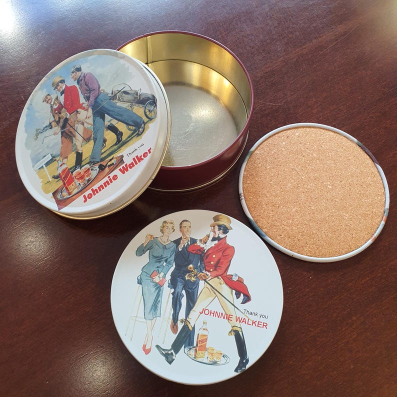 May include: A vintage Johnnie Walker tin with a lid featuring a golf scene and two coasters. One coaster has a cork surface, and the other has a Johnnie Walker illustration. The tin is open, revealing its interior.