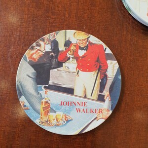 May include: Round coaster featuring a vintage illustration with the text "JOHNNIE WALKER." The image depicts several men in a room, with a man in a red coat and a bottle of whiskey. Two glasses of whiskey are on a table.