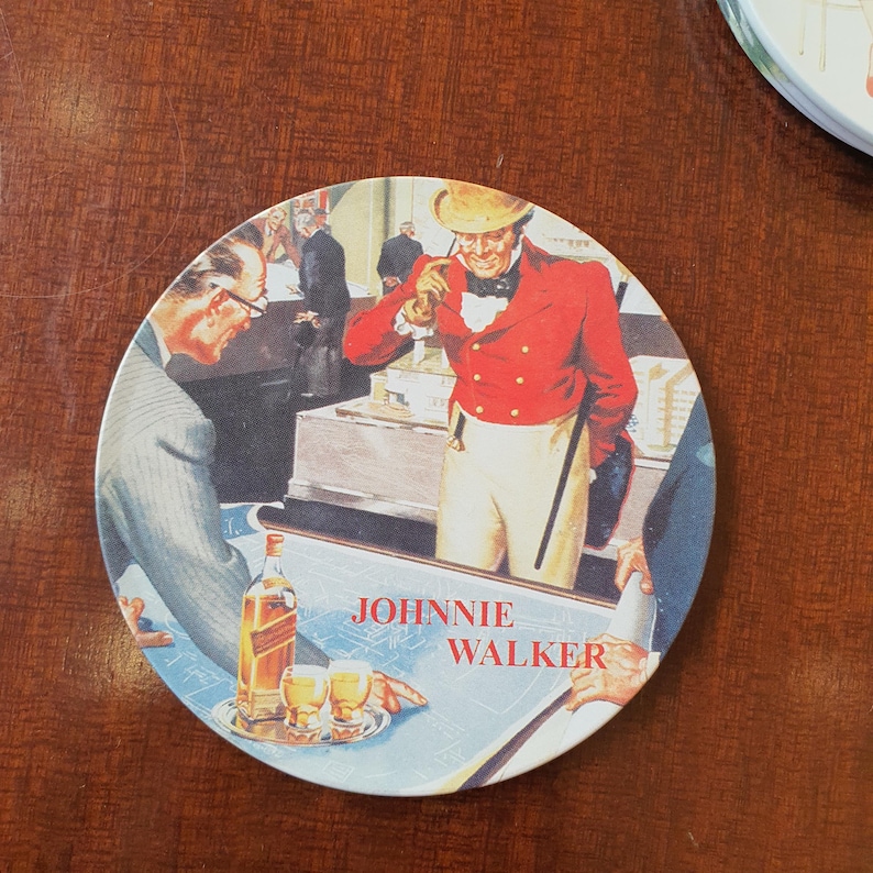 May include: Round coaster featuring a vintage illustration with the text "JOHNNIE WALKER." The image depicts several men in a room, with a man in a red coat and a bottle of whiskey. Two glasses of whiskey are on a table.