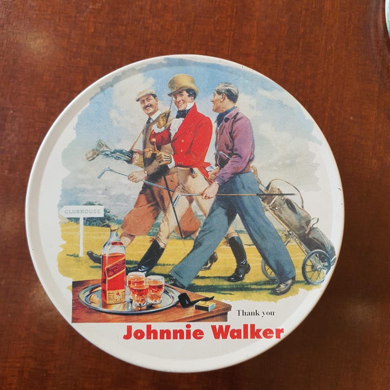 May include: Round metal tray with a vintage illustration of three golfers walking on a green. A bottle of Johnnie Walker whiskey, glasses, and text are featured. The tray has a white border and a brown wooden background.