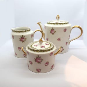 Könnte beinhalten: Ein Vintage-Teeservice mit Teekanne, Sahnekännchen und Zuckerdose. Die weißen Porzellanstücke sind mit rosa Rosenmustern und goldenen Akzenten verziert. Die Teekanne hat einen Deckel und einen gebogenen Griff. Die Zuckerdose hat einen Deckel und zwei Griffe.
