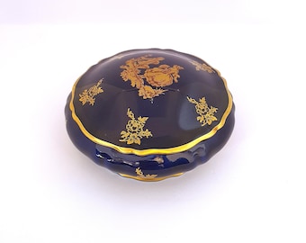May include: A navy blue, ceramic trinket box with a scalloped edge and gold trim. The lid features gold floral and ornate designs. The box is round and sits on a white surface.