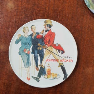 May include: Round white decorative plate featuring a vintage illustration of three people and a man in a red coat, with the text "Thank you JOHNNIE WALKER". The plate has a glossy finish and a brown rim.