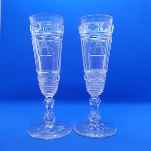 May include: Two clear, etched champagne flutes with a detailed design. Each glass features a wide base, a slender stem, and a bowl with an etched monogram. The rims have a decorative pattern. The background is a solid blue.