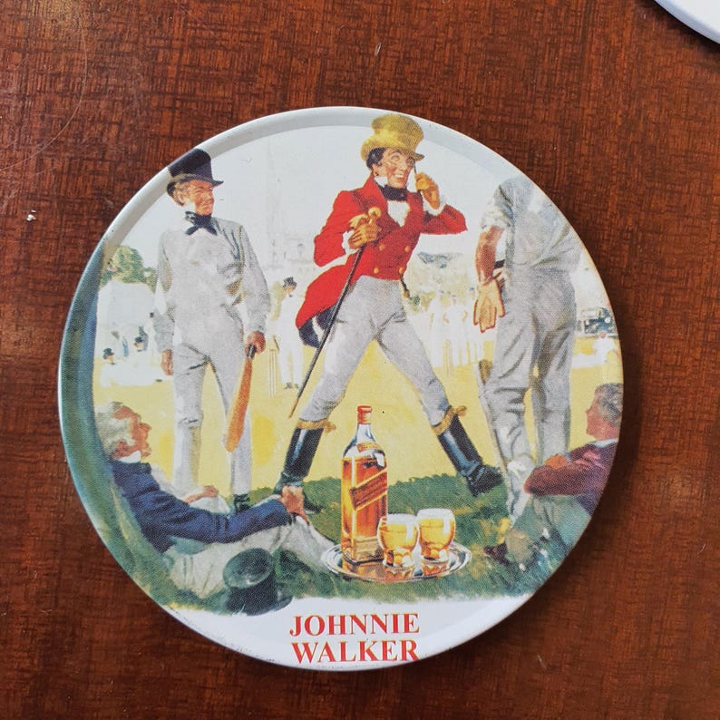May include: Round coaster with a vintage illustration of people at a cricket match. A bottle of Johnnie Walker whiskey and two glasses are on a tray. The coaster has the words "JOHNNIE WALKER" in red.